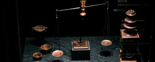 A set of copper or bronze balance-style scales with one end higher than the other, on a dark tabletop with small cups and other paraphernalia scattered about